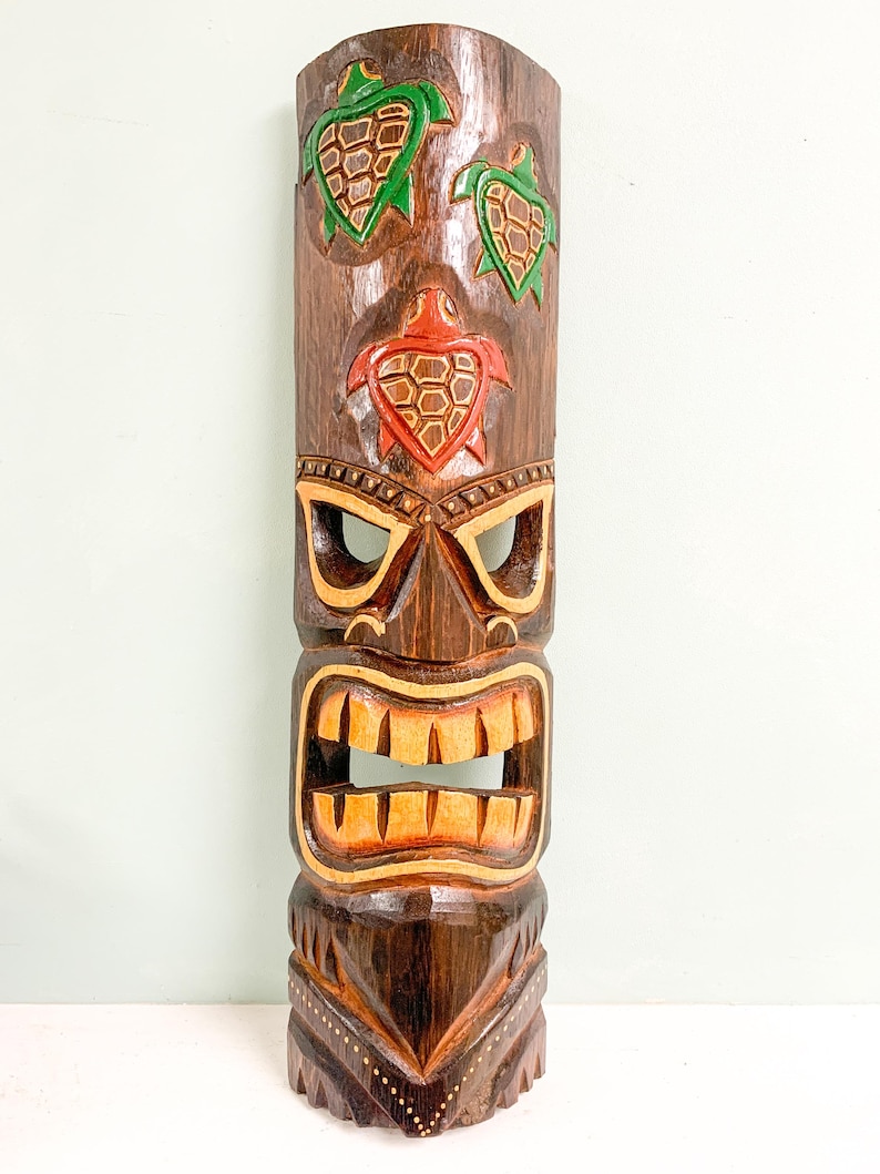 May include: A wooden tiki mask with a carved face and three green and red turtle designs. The mask is brown with yellow accents.