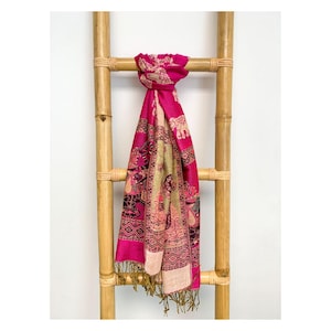 May include: A bright pink scarf with an elephant and floral pattern, displayed on a bamboo ladder. The scarf has a mix of pink, gold, and cream colours, and a fringed edge. The scarf is approximately 180cm in length.