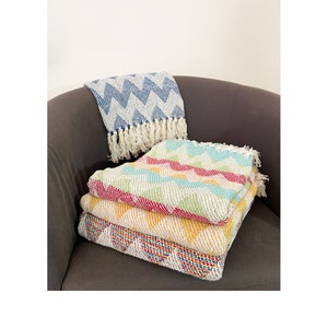 May include: A stack of four colourful woven throw blankets with geometric patterns in shades of blue, green, red, yellow, and white. The blankets are folded and stacked on a grey armchair.