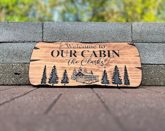 May include: Wooden sign with the text "Welcome to Our Cabin The Clarks" and a cabin illustration surrounded by trees.