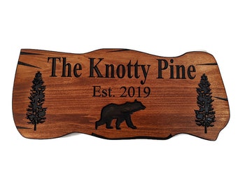 May include: A wooden sign with a rustic, irregular shape. The sign is brown with black lettering and a silhouette of a bear and two pine trees. The text reads "The Knotty Pine Est. 2019".