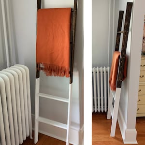 May include: A white ladder shelf with brown wooden accents, painted white, with an orange throw blanket draped over the top rung. The ladder shelf is standing in a room with white walls and a white radiator.