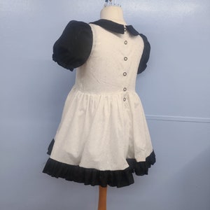 Pu&ograve; includere: Un vestito bianco con maniche a sbuffo nere e un bordo di volant nero. Il vestito ha un colletto Peter Pan nero e una fila di bottoni neri sul retro.