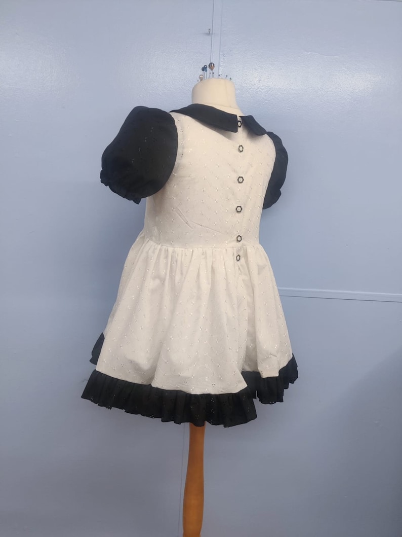 Pu&ograve; includere: Un vestito bianco con maniche a sbuffo nere e un bordo di volant nero. Il vestito ha un colletto Peter Pan nero e una fila di bottoni neri sul retro.