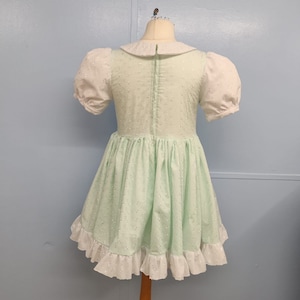 Pu&ograve; includere: Un vestito verde chiaro con colletto bianco e maniche a sbuffo. Il vestito ha un bordo di pizzo bianco intorno al fondo e una cerniera sul retro.