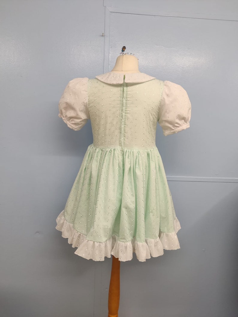 Pu&ograve; includere: Un vestito verde chiaro con colletto bianco e maniche a sbuffo. Il vestito ha un bordo di pizzo bianco intorno al fondo e una cerniera sul retro.