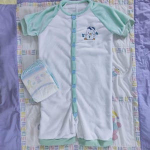 Peut inclure: Une grenouillère blanche pour bébé avec une bordure verte et un petit motif de pingouin brodé. La grenouillère a une fermeture boutonnée sur le devant.