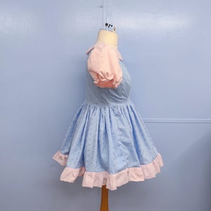 May include: A light blue dress with a pink collar and puffed sleeves. The dress has a white eyelet pattern and a pink ruffle trim.