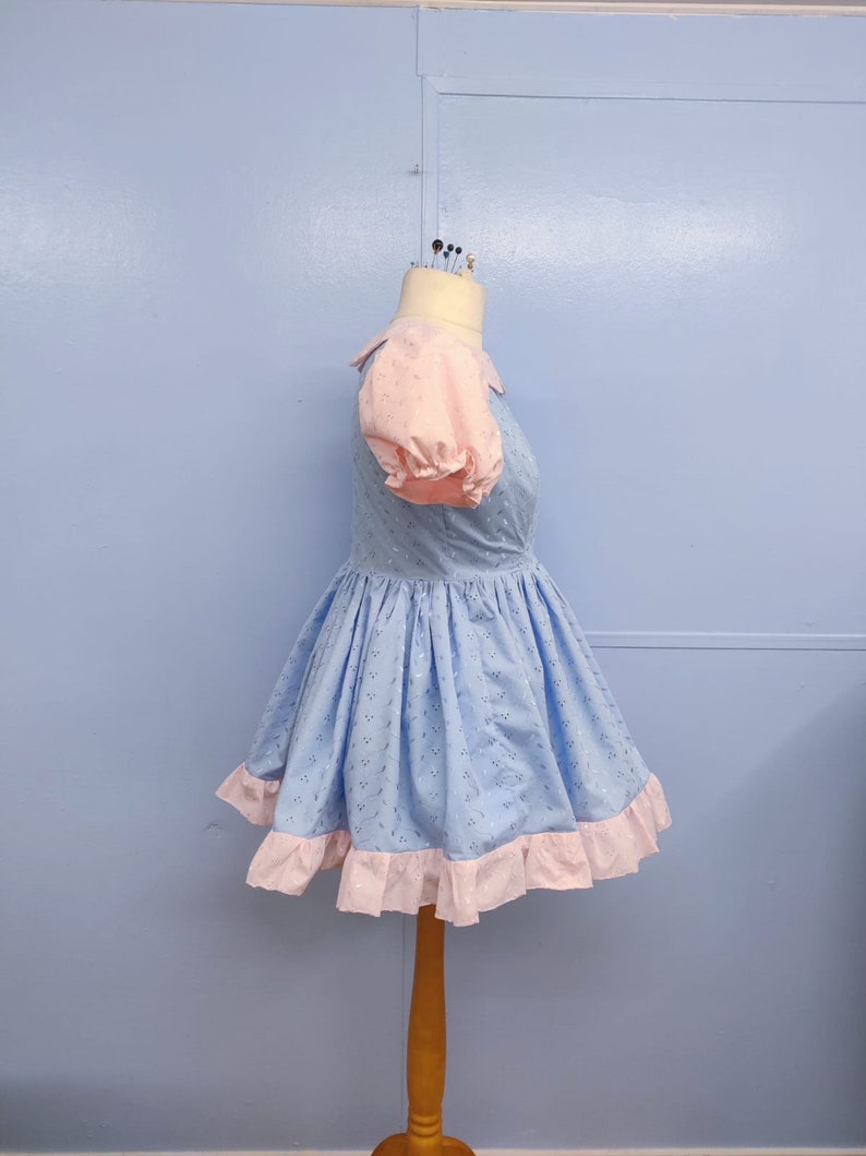 May include: A light blue dress with a pink collar and puffed sleeves. The dress has a white eyelet pattern and a pink ruffle trim.