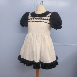 Pu&ograve; includere: Un vestito bianco con bordi neri e un colletto nero. Il vestito ha un motivo a pois e un orlo a balze.