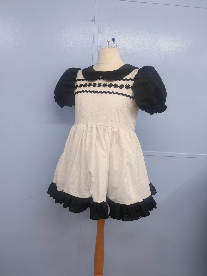 Pu&ograve; includere: Un vestito bianco con bordi neri e un colletto nero. Il vestito ha un motivo a pois e un orlo a balze.