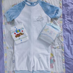 Könnte beinhalten: Ein weißer und blauer Baby-Schlafsack mit blau-weißem Streifenbesatz. Der Schlafsack hat eine blaue und weiße gestickte Wolke mit Mond und Sternen auf der Vorderseite. Der Schlafsack liegt auf einer lila und weißen Steppdecke mit einem bunten Rand. Auf der Steppdecke liegen zwei Pakete mit Babywindeln.
