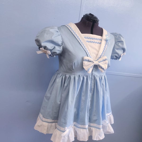 May include: A light blue dress with a sailor collar design. The dress features a white collar with a wavy trim, a matching bow, and puff sleeves. The skirt has a ruffled hem with white accents and bows. The dress is displayed on a mannequin.