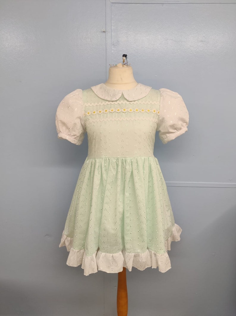 Pu&ograve; includere: Un abito verde chiaro con colletto bianco e maniche a sbuffo bianche. L'abito &egrave; rifinito con un bordo in pizzo bianco con ricami di margherite gialle. La gonna &egrave; a trapezio e ha un volant in pizzo bianco sull'orlo.