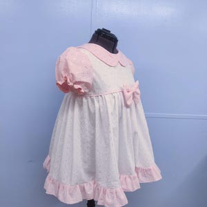 Puede incluir: Un vestido blanco con ribetes rosas y un lazo rosa. El vestido tiene un fondo blanco con pequeñas flores rosas y un cuello rosa.