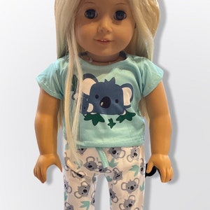 May include: A doll wearing a light blue t-shirt with a koala print and white pants with a koala print. The doll has blonde hair and blue eyes.