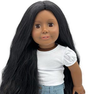 May include: A doll with long black hair, brown eyes, and a smiling expression. The doll is wearing a white short-sleeved shirt and a denim skirt with frayed edges. The doll is standing upright.