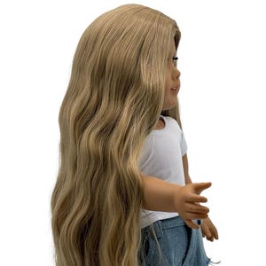 May include: A doll with long, wavy, light brown hair. The doll is wearing a white t-shirt and denim shorts. The doll is standing and facing to the side.