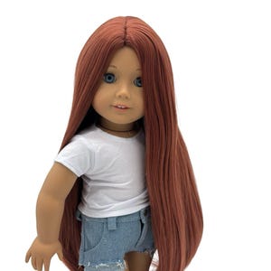 Puede incluir: Una muñeca con pelo largo color castaño rojizo, ojos azules y una camiseta blanca. La muñeca lleva pantalones cortos vaqueros. La muñeca está de pie y mirando hacia delante.