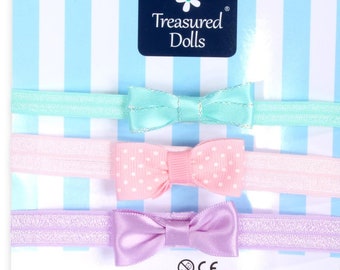 May include: A set of three hair ties with bows. The bows are in different colours and patterns: light blue, pink with white polka dots, and light purple. The hair ties are packaged on a white and blue striped background with the text "Treasured Dolls" in a blue box.