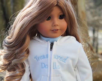 May include: A doll wearing a white hoodie with the text "Treasured Dolls" embroidered on the front. The hoodie has a blue zipper and a blue flower embroidered on the pocket. The doll is wearing blue jeans.