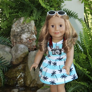 May include: A doll wearing a blue and white striped dress with palm trees printed on it. The doll is wearing white sandals and sunglasses. The doll is holding a small camera around her neck.