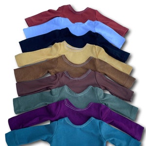 K&ouml;nnte beinhalten: Ein Stapel von zehn lang&auml;rmeligen Hemden in verschiedenen Farben, darunter Rot, Blau, Schwarz, Gelb, Braun, Gr&uuml;n, Lila und T&uuml;rkis.