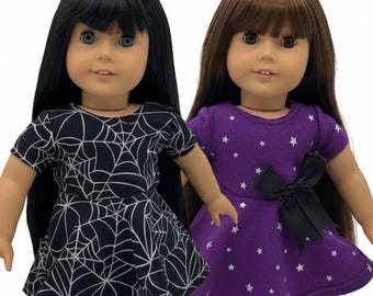 May include: Two dolls with long hair and blue eyes. One doll wears a black dress with a white spiderweb pattern, the other a purple dress with white stars and a black bow. Both dolls wear black shoes with heart-shaped embellishments.