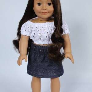 May include: A doll with long brown hair wearing a white eyelet top and a denim skirt with glitter. The doll is wearing white sandals.
