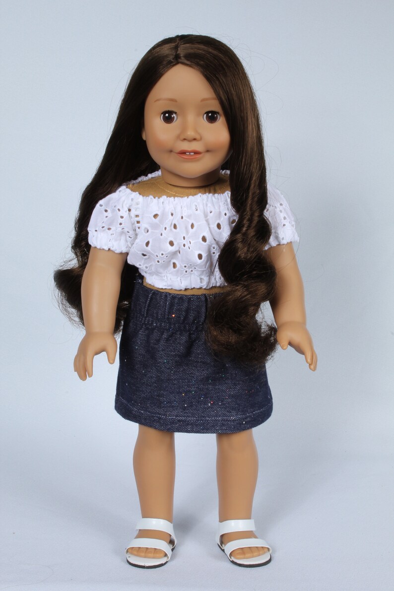 May include: A doll with long brown hair wearing a white eyelet top and a denim skirt with glitter. The doll is wearing white sandals.
