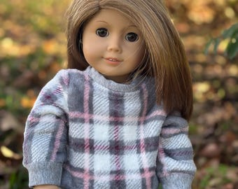 May include: A doll with long brown hair wearing a gray and pink plaid sweater and pink shorts. The doll is standing in front of a blurred background of fall leaves.