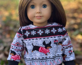 Puede incluir: Una muñeca con un suéter navideño gris y negro con un patrón de dachshund. El suéter tiene rayas de bastón de caramelo rojo y blanco y copos de nieve. La muñeca tiene el pelo largo y castaño y ojos azules.