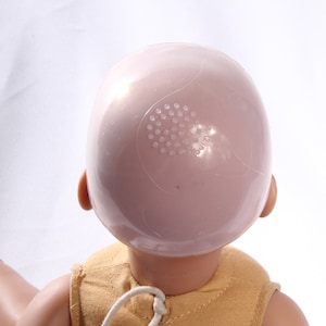May include: A doll head with a pink plastic cap on top. The cap has a small hole with a grid pattern and the word "build-a-bear" printed on it.