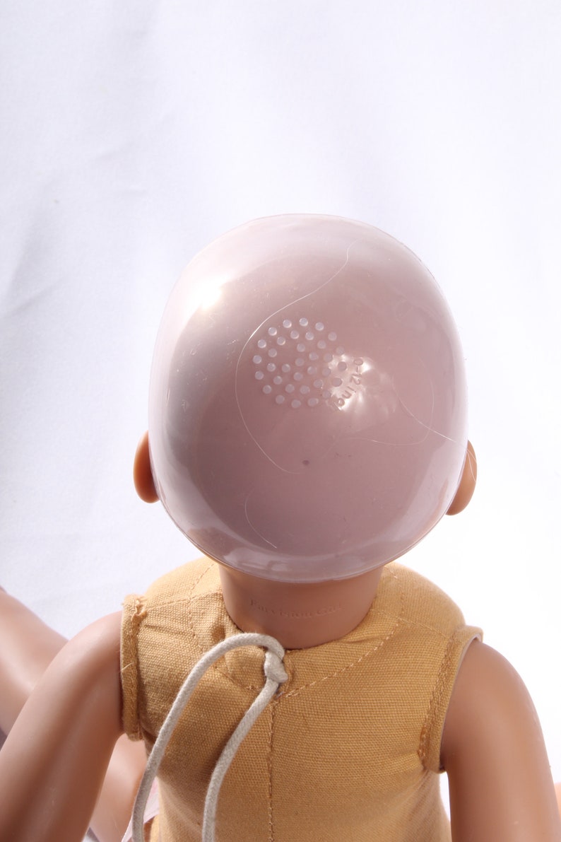 May include: A doll head with a pink plastic cap on top. The cap has a small hole with a grid pattern and the word "build-a-bear" printed on it.