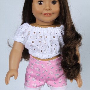 May include: A doll wearing a white eyelet crop top and pink floral print shorts.