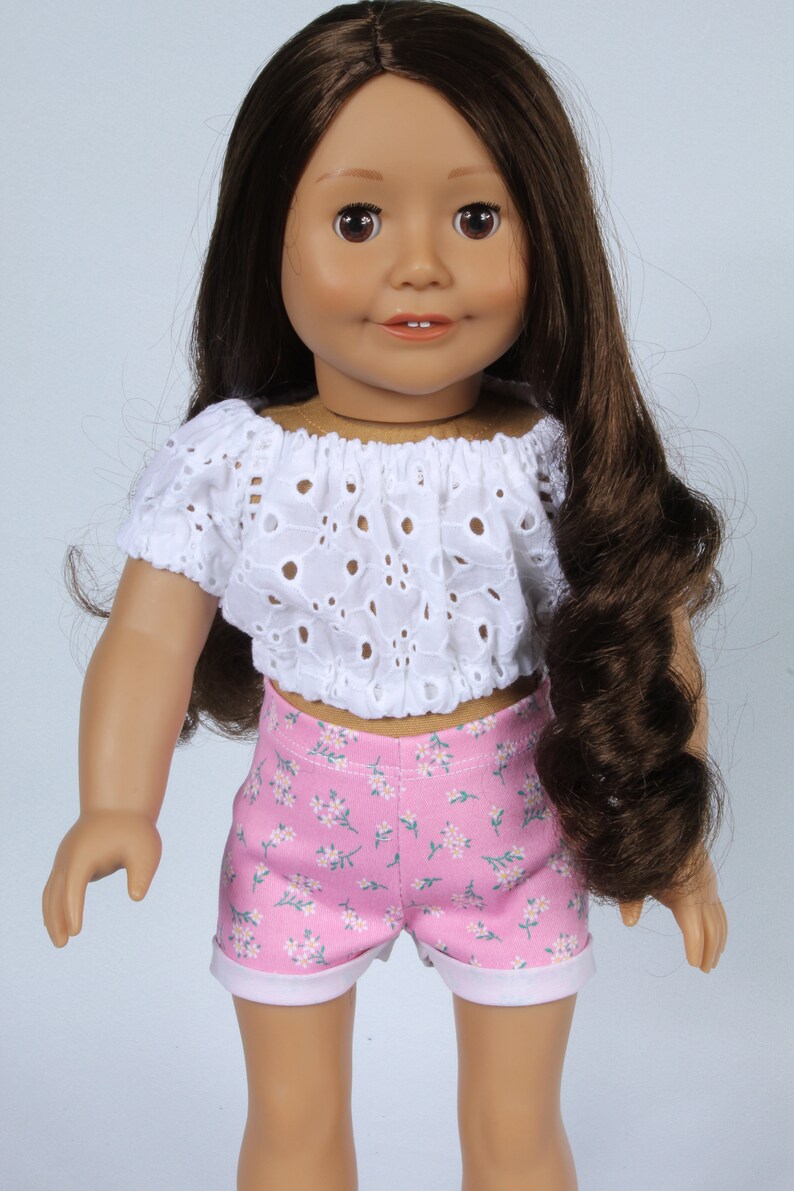 May include: A doll wearing a white eyelet crop top and pink floral print shorts.