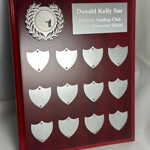 May include: A dark wood plaque featuring a silver circular emblem with a fishing silhouette, a silver rectangular plate engraved with "Donald Kelly Snr Kintyre Angling Club Junior Memorial Shield," and twelve small silver shield-shaped plates.