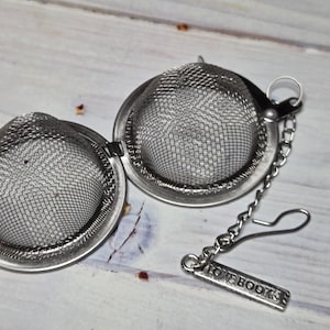 May include: A stainless steel tea infuser with a mesh basket design. The infuser is open, revealing the interior. A chain and hook are attached, with a rectangular charm that reads "LOVE BOOKS".