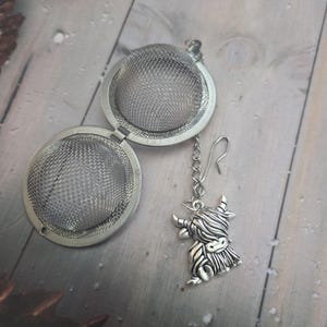 May include: A silver tea infuser with a hinged, mesh ball design. A chain and hook are attached, with a Highland cow charm. The infuser is open, showing the mesh interior. The charm is detailed with the cow's features.