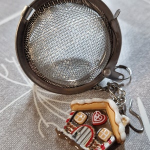 May include: A silver tea infuser with a mesh ball and a gingerbread house charm. The gingerbread house is decorated with white icing, red candy hearts, and brown candy canes.