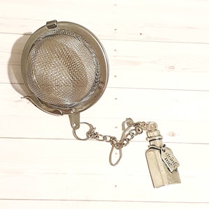 May include: A stainless steel tea infuser with a mesh basket and a chain attached to a bottle charm. The charm has the words "drink me" on a small tag. The infuser is designed for steeping loose leaf tea.