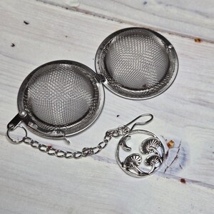 May include: A stainless steel tea infuser with a chain and a decorative charm featuring mushroom designs. The infuser is open, revealing the mesh interior, and rests on a light wood surface.