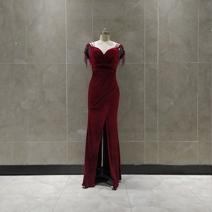 May include: A long, burgundy velvet dress with a sweetheart neckline and beaded straps. The dress has a fitted bodice and a flowing skirt with a slit in the front.