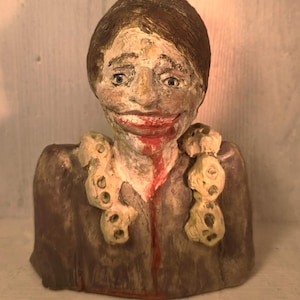 May include: A handmade ceramic bust with a disturbing expression. The figure has brown hair, blue eyes, and a wide, unsettling smile with red lipstick. A red streak runs down the chest. The figure wears a brown garment with cream-colored embellishments.