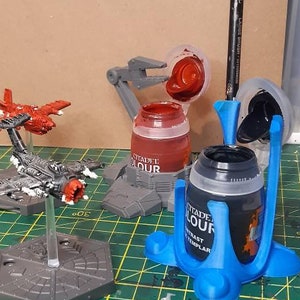 May include: Two miniature spaceships on grey bases, one red and one grey, are on a green cutting mat. Two paint pots, one red and one black, are on a blue paint pot holder. A paintbrush is in the black paint pot holder.