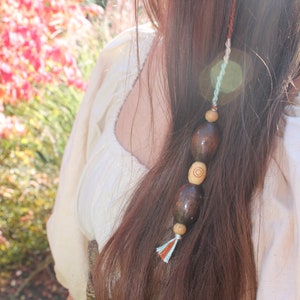 May include: A brown hair tie with wooden beads and a blue, white, and orange tassel. The hair tie is tied into a woman's long brown hair.