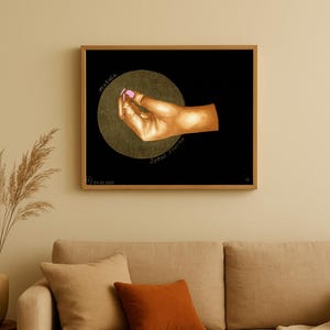 Può includere: Un'opera d'arte incorniciata raffigurante una mano con smalto rosa contro un cerchio dorato su sfondo nero. Le parole "Miracle" e "Spout Power" sono scritte attorno al cerchio. L'opera è sopra un divano beige con cuscini arancioni e beige.