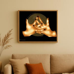 Può includere: Un'opera d'arte incorniciata con un'illustrazione dettagliata di due mani in un gesto di fiducia. Le mani sono raffigurate con una calda tonalità dorata su uno sfondo nero, con le parole "Vajrapradma" e "Trust" visibili. L'opera d'arte è esposta sopra un divano beige.