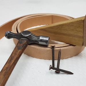 May include: A close-up of leatherworking tools, including a hammer, a wooden wedge, a leather strap, and three metal nails.