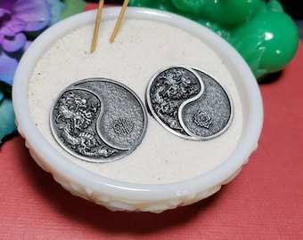 May include: A white ceramic bowl filled with sand and two silver yin yang charms with dragon designs. The bowl is sitting on a red surface with two incense sticks in the background.